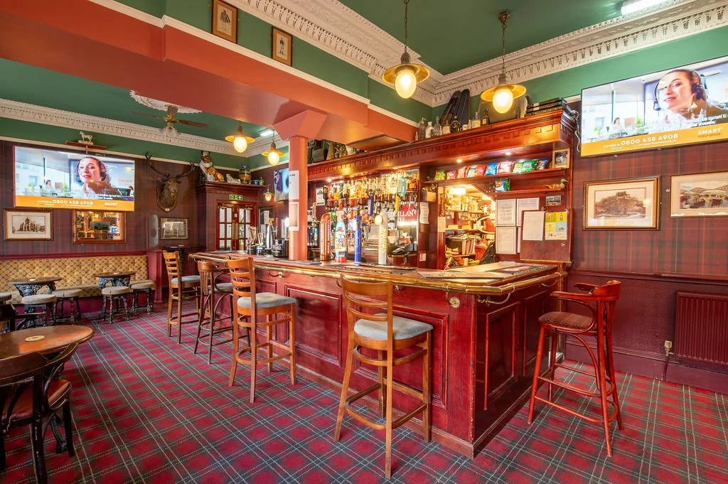 This is a cozy pub or bar with wooden furniture, a tiled tartan patterned carpet, and framed pictures on the walls. There are multiple television screens displaying a woman with headphones. The bar counter has seating and is stocked with bottles and 