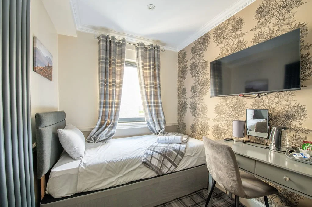 A bedroom with a single bed, gray upholstered headboard, white bedding, and a plaid blanket. A window with plaid curtains is behind the bed. Opposite the bed, a wall-mounted flat-screen TV is above a white desk with a mirror, a lamp, kettle, and tray