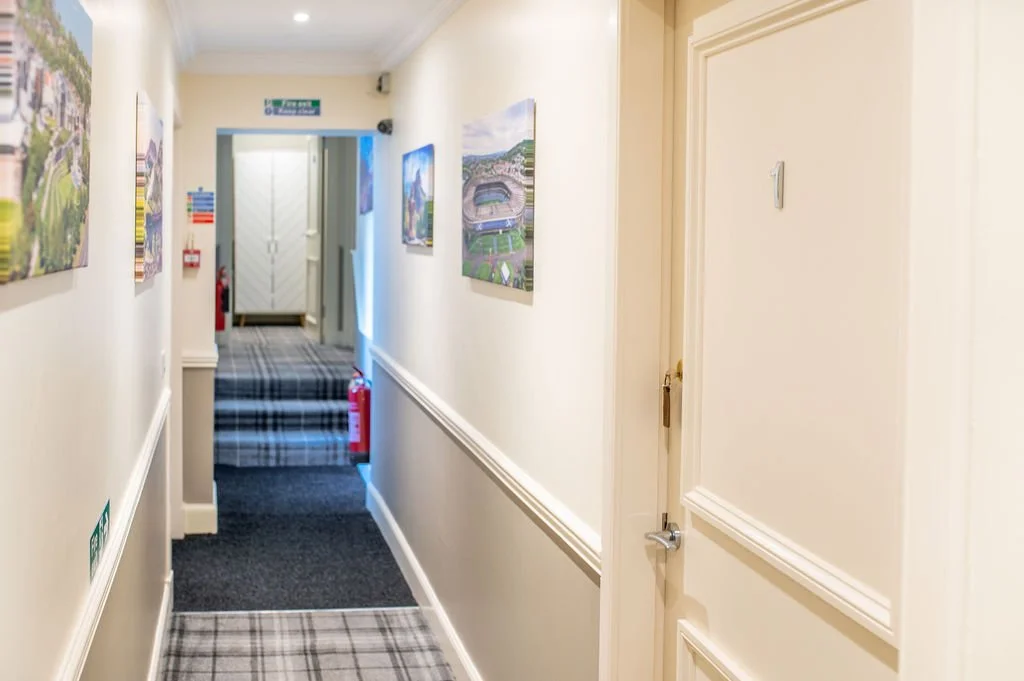 Hallway with framed pictures, a staircase with a plaid carpet, fire extinguisher, and an emergency exit sign.