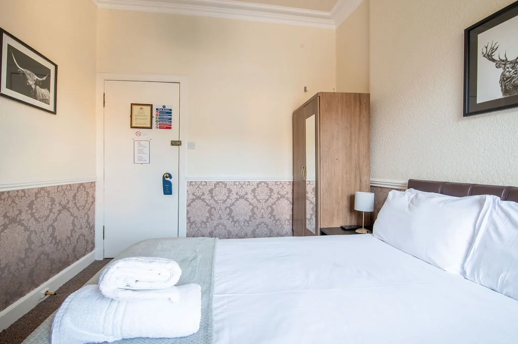 Hotel room with a bed, white linens, a wooden wardrobe, a bedside table with a lamp, and framed deer artwork on the walls.