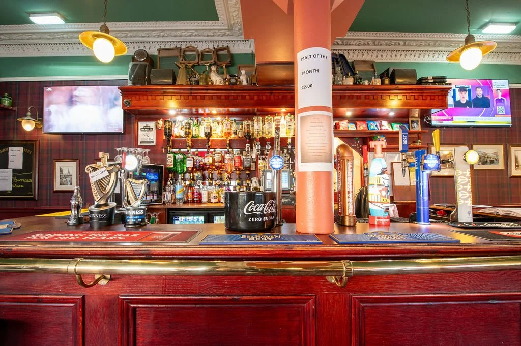 A bar with a wooden counter and various alcohol bottles behind it, along with beer taps, beer mats, and a Coke Zero sugar bucket. There are two television screens and framed pictures on the wall, with a plaid wallpaper background.