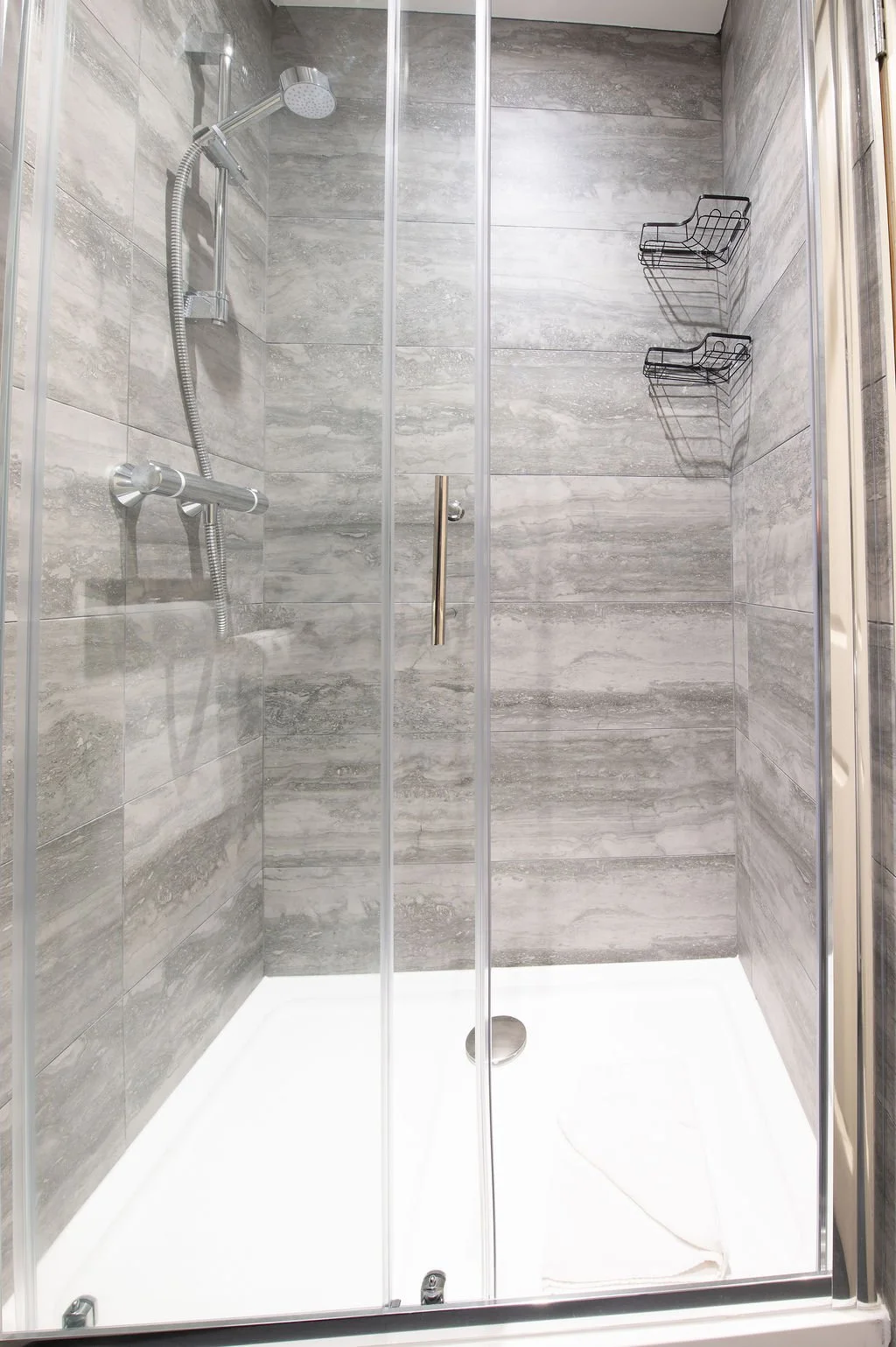 A modern shower stall with gray tiled walls, a glass door, a handheld showerhead, and black wire shelves on the wall.