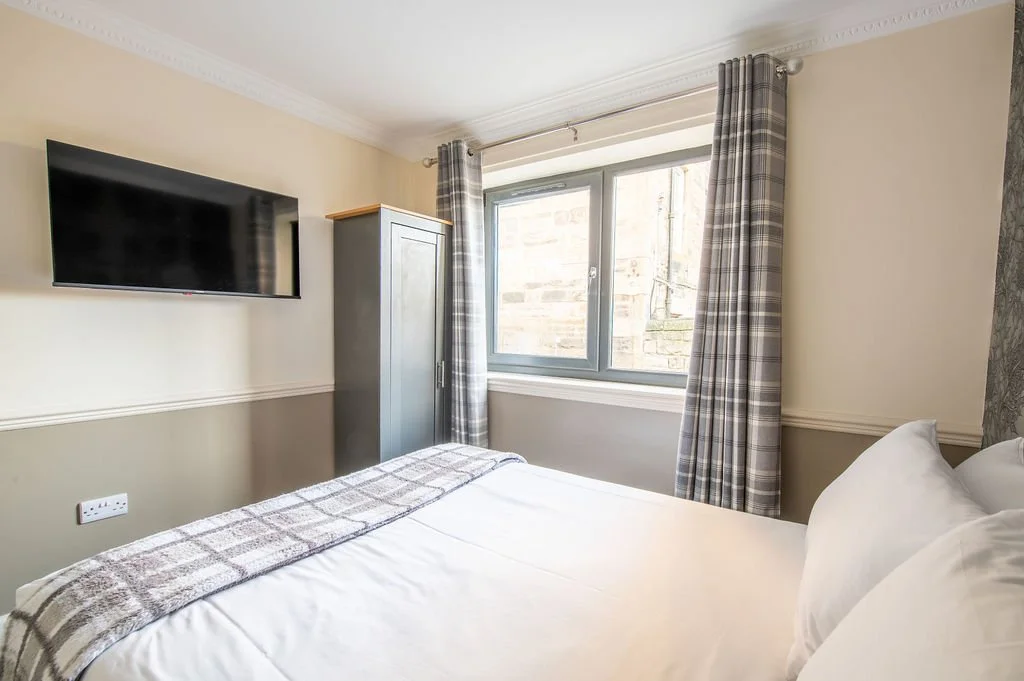 A hotel room with a bed, a flat-screen TV on the wall, a wardrobe, a large window with plaid curtains, and a light-colored wall.