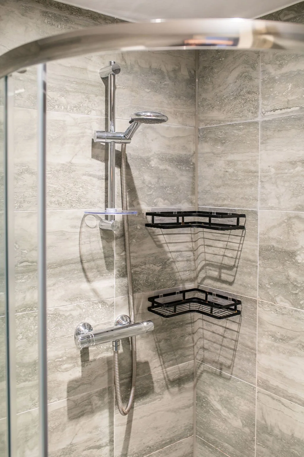 A close-up view of a modern shower with gray stone tile walls, a chrome adjustable showerhead, and two black wire corner shelves.