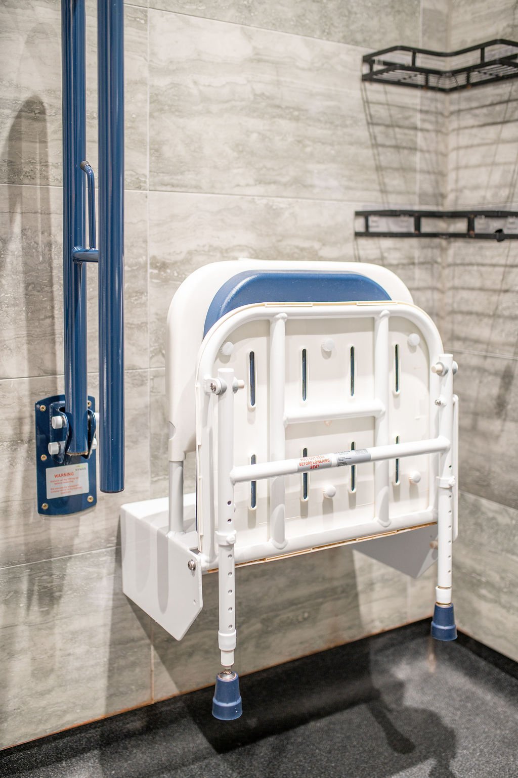 Hospital shower with a fold-down chair, blue grab bar, and wall-mounted shelves.