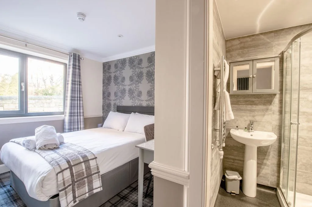 A bedroom with a large bed, decorated with neutral tones and plaid bedding, next to a window with curtains, and an adjoining modern bathroom with a pedestal sink, mirror cabinet, and glass shower enclosure.