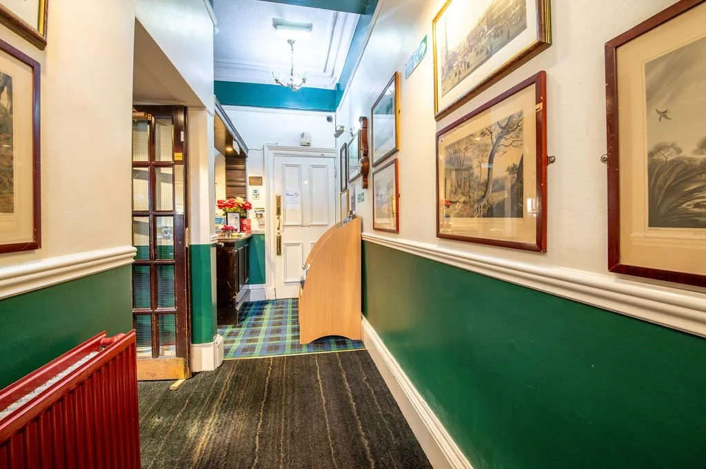 Hallway inside a building decorated with framed artwork and painted green and cream walls, with a small area near the entrance showing a black tiled floor and some red flowers.