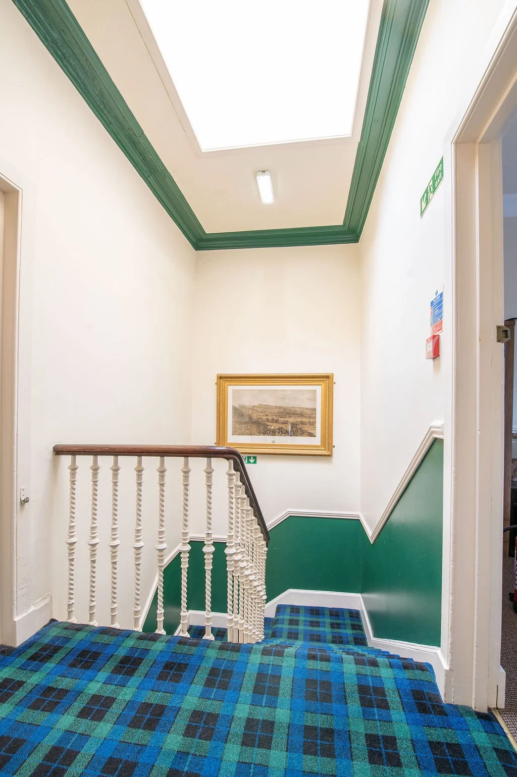 Interior view of a stairwell landing showing a patterned carpet, painted green and white walls, a framed picture, and a ceiling with a light and a skylight.