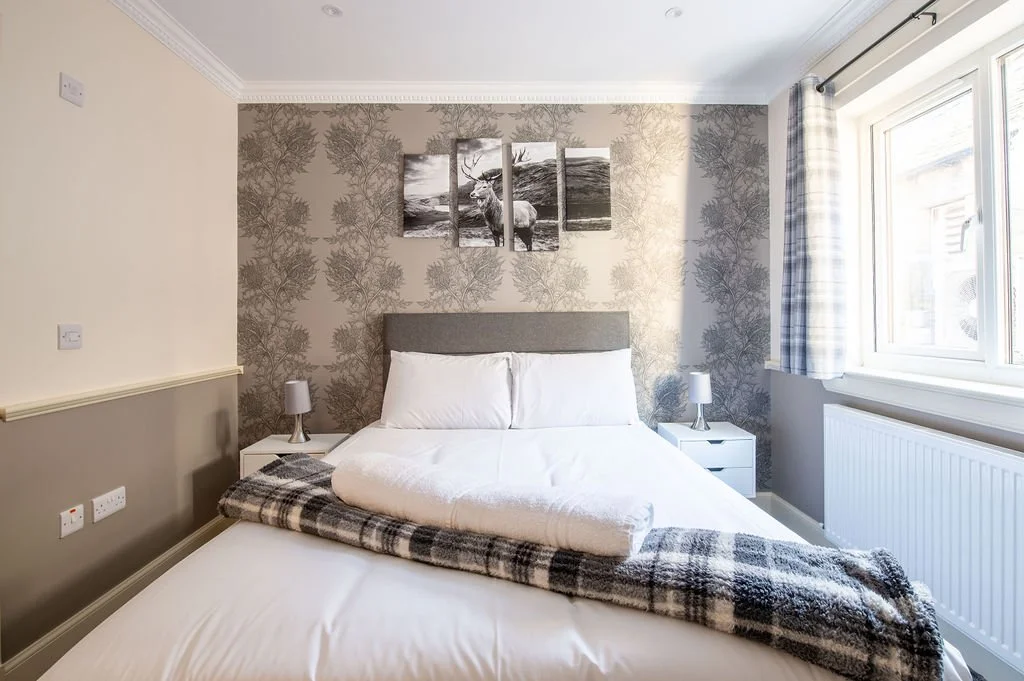 A bedroom with a bed, pillows, blanket, two nightstands with lamps, patterned wallpaper on the back wall, and a window with curtains.