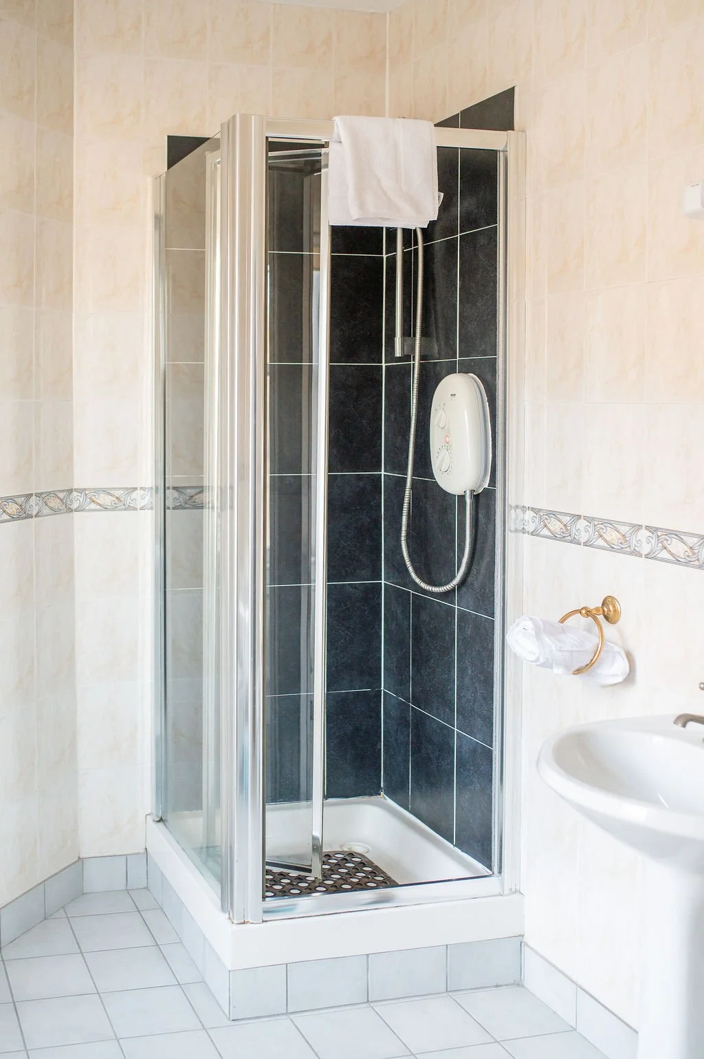 A corner shower with black and beige tiled walls, a white towel hanging on top, a white electric shower panel, and a small white sink nearby.