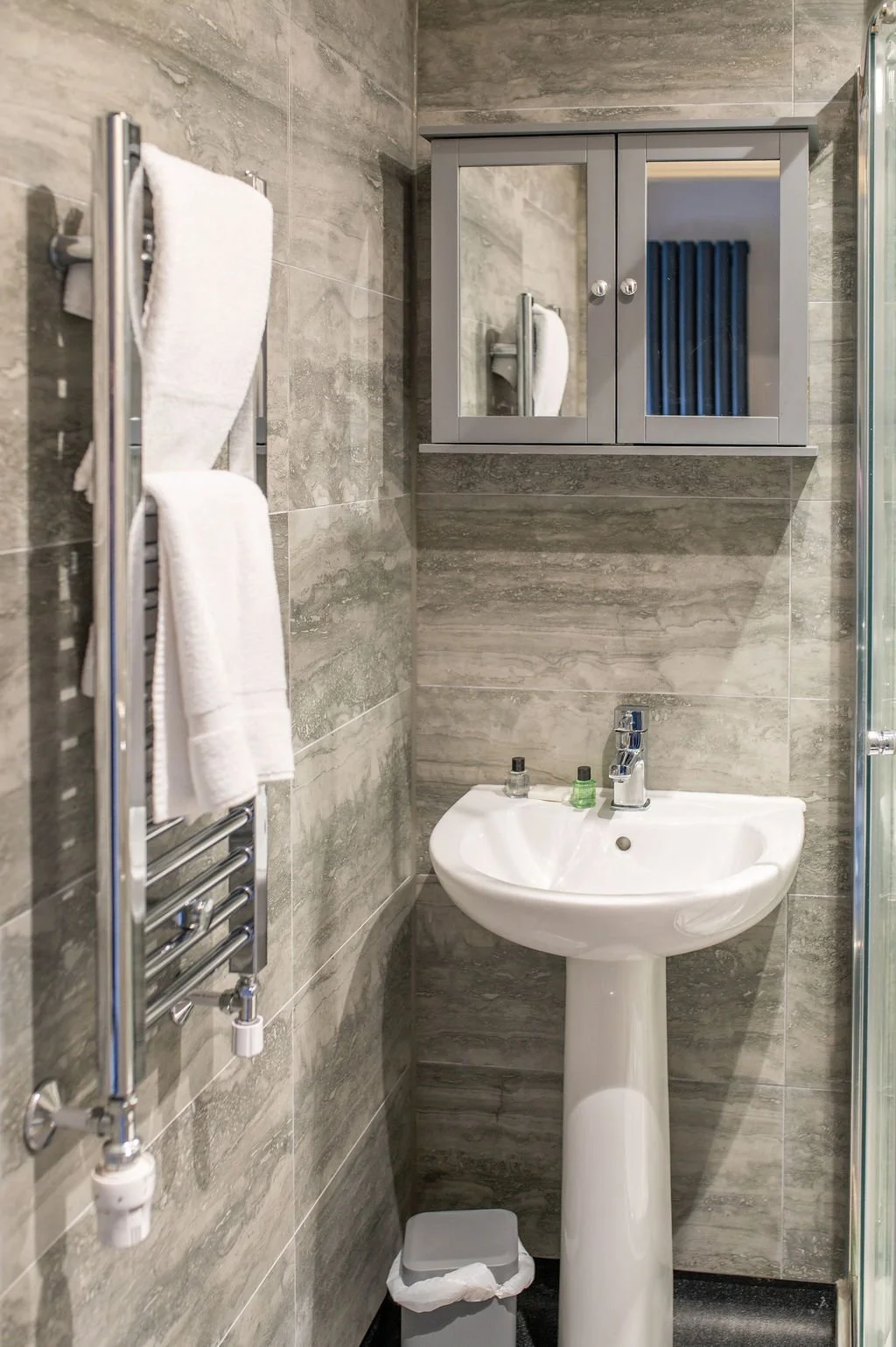 A small bathroom with gray tiled walls, a white pedestal sink with a faucet and soap dispensers, a mirror cabinet above the sink, and a wall-mounted towel radiator with white towels. There is a small trash can beneath the sink.