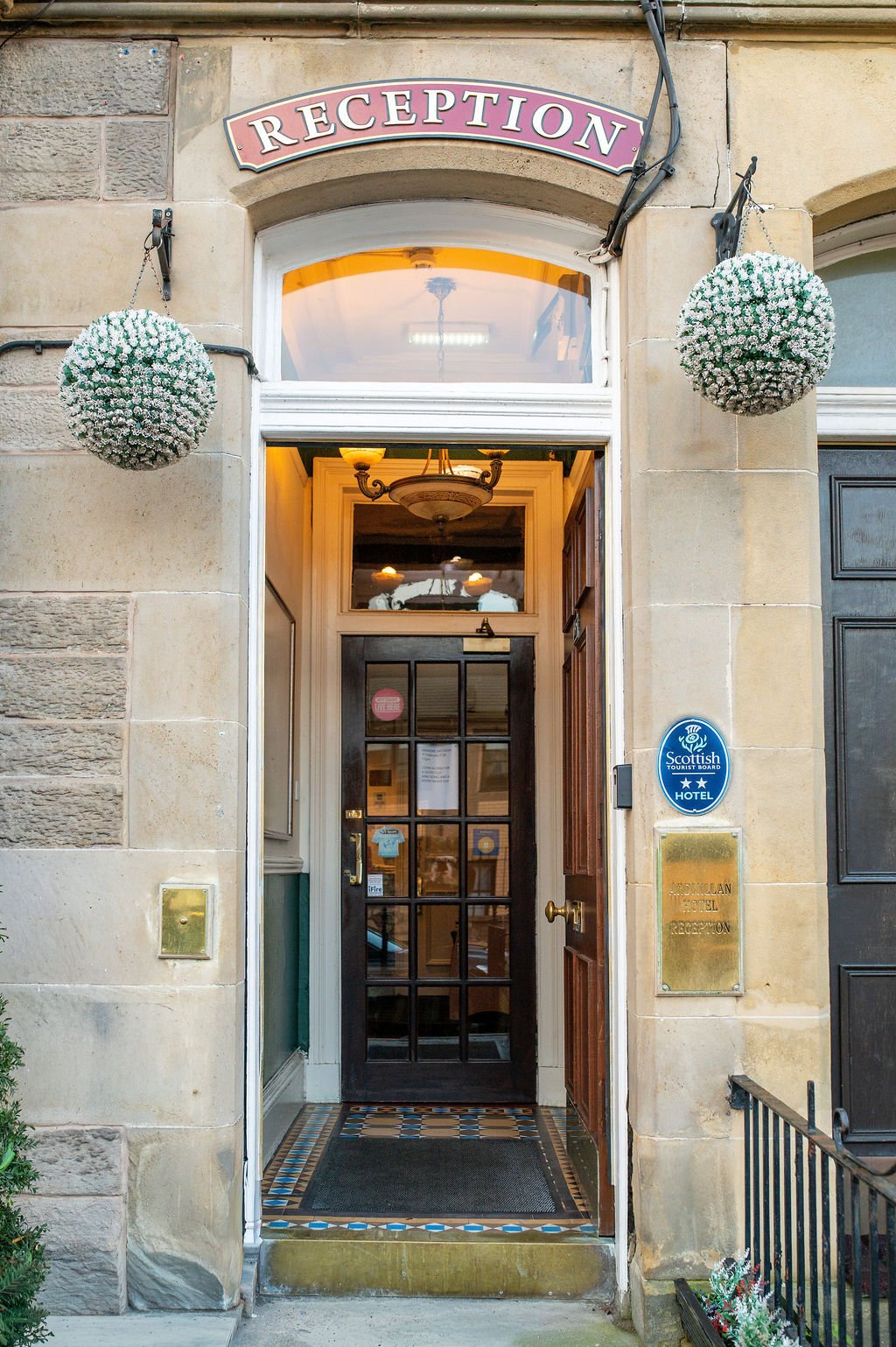 Entrance to a hotel with a sign reading 'RECEPTION' above the door, two hanging white globe-shaped flowers, plaque indicating Scottish Tourist Board hotel, and a black door with glass panes inside a stone building.
