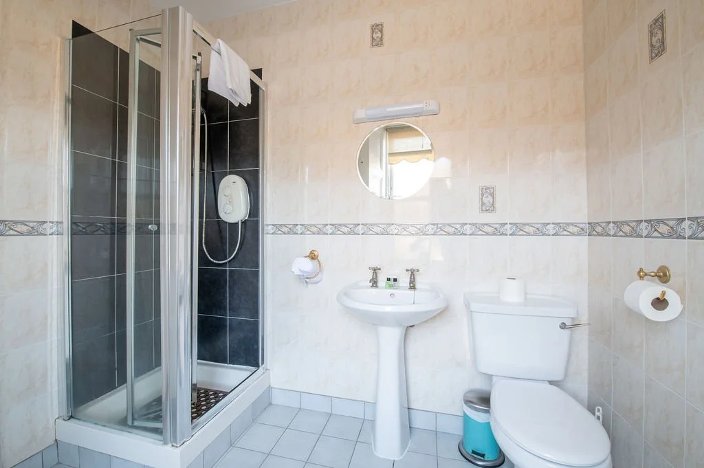 Bathroom with a shower stall, a pedestal sink, a toilet, and wall-mounted fixtures, beige tiled walls with decorative border, and a round mirror above the sink.