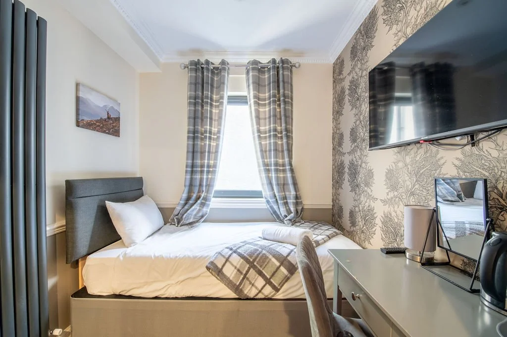 A cozy hotel room with a bed, patterned curtains, a wall-mounted TV, a small desk with a mirror, lamp, and kettle, and a framed picture on the wall.