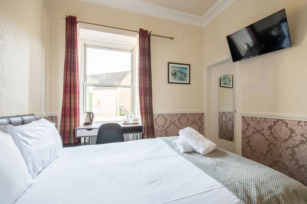A hotel room with a bed, a desk with a chair, a window with curtains, a wall-mounted TV, and framed artwork.