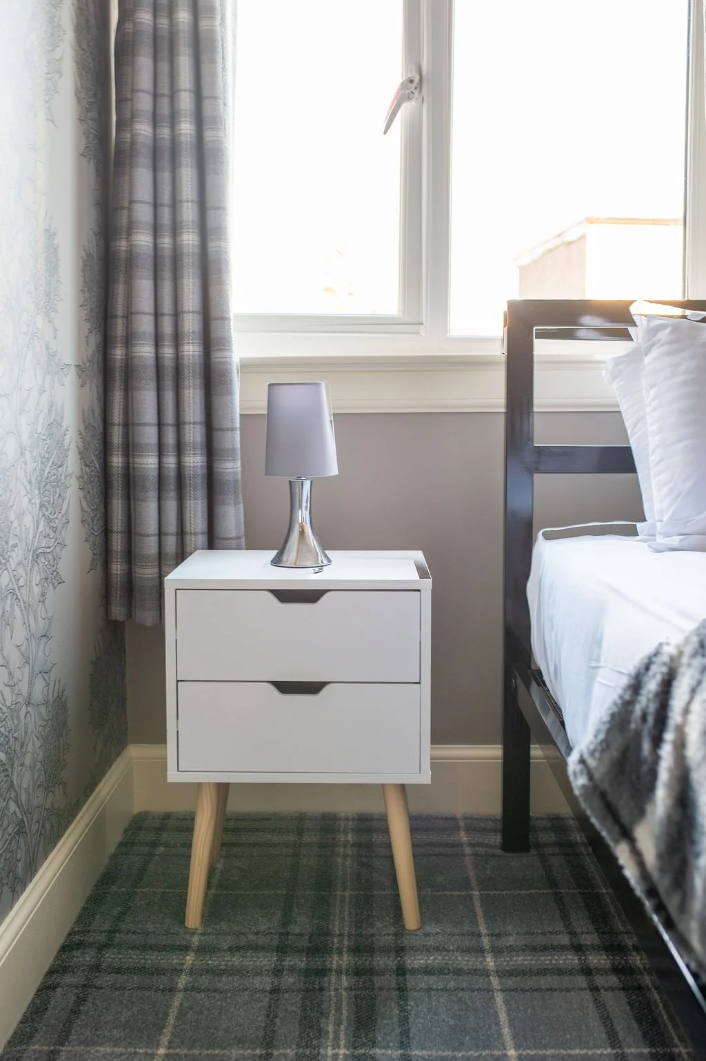 A white bedside table with two drawers and wooden legs, with a silver table lamp on top, positioned next to a bed with white bedding, in front of a window with plaid curtains.