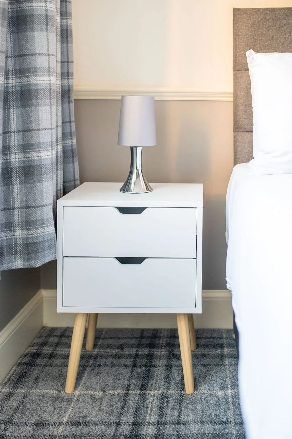 A white bedside table with two drawers, a metallic lamp with a white lampshade on top, next to a bed with white bedding, in a hotel room with plaid curtains and a grey and black checkered carpet.
