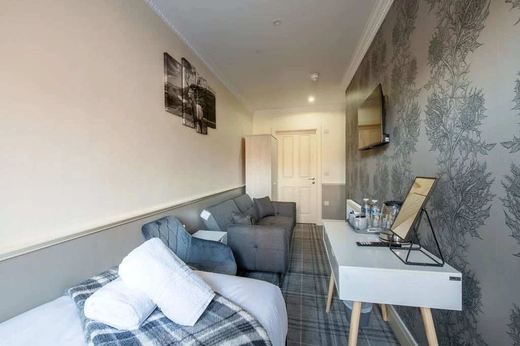 Hotel room with bed, sofa, wall-mounted TV, and a white desk with water bottles and tea set.