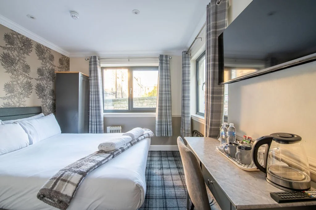Hotel room with a bed, wardrobe, window with plaid curtains, and a desk with bottled water, cups, a coffee maker, and a TV on the wall.