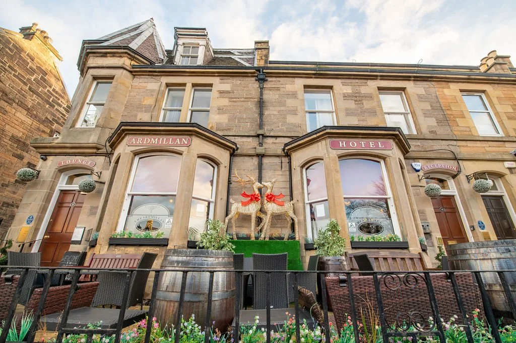 Exterior view of The Ardmillan Hotel, a historic stone building with large bay windows, decorative reindeer statues with red bows, surrounded by barrels, plants, and outdoor seating.