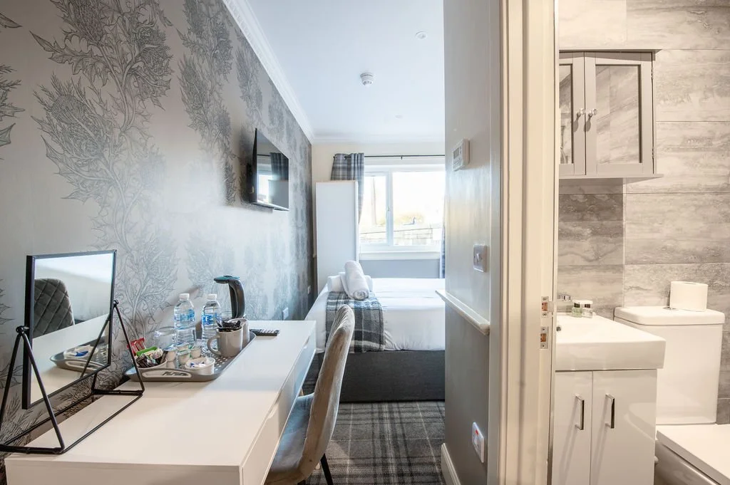 A hotel room with a bed near a window, a desk with bottles of water, a mirror, and a tissue box, and an adjacent bathroom with a sink and toilet.