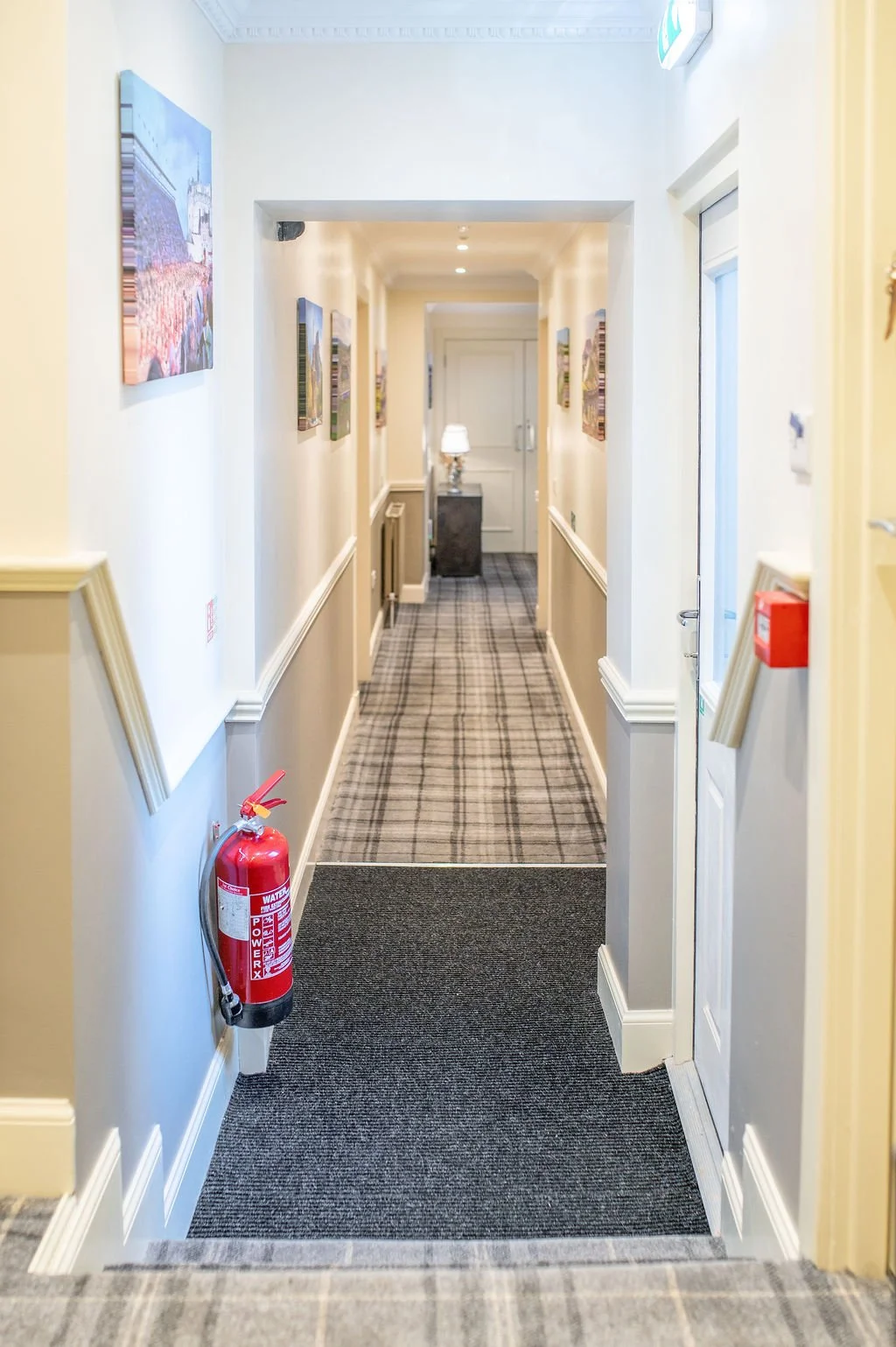 Long hotel corridor with cream-colored walls, framed artwork, patterned carpet, and a table with a lamp at the far end.