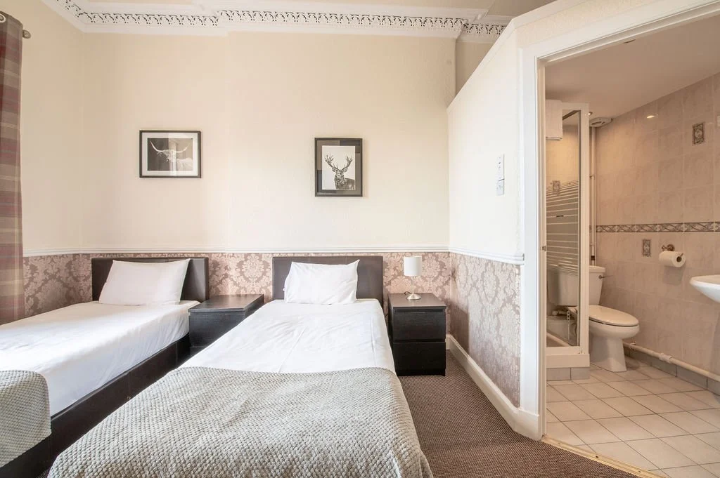 A hotel room with two single beds, a nightstand, a lamp, framed pictures of animals on the wall, and a visible bathroom with a toilet, shower, and tiled walls.