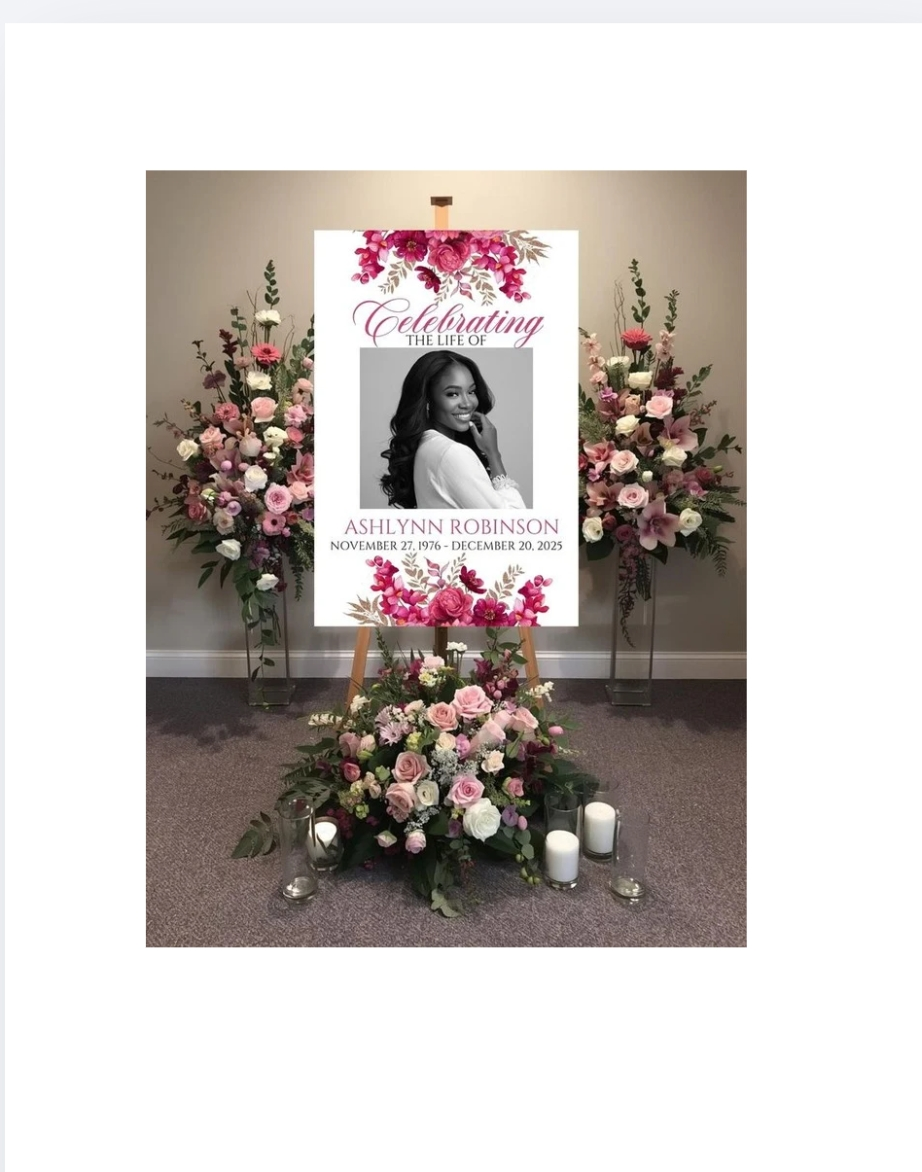 Funeral or memorial display with pink and white flowers, candles, and a poster honoring Ashlynn Robinson, who lived from November 27, 1976, to December 20, 2025, featuring a photo of her smiling.