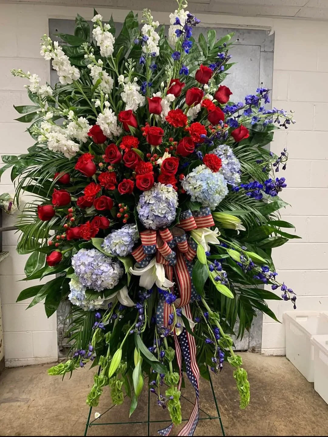 A large, colorful floral arrangement with red roses, white lilies, purple delphiniums, and blue hydrangeas, decorated with patriotic ribbons in red, white, and blue.