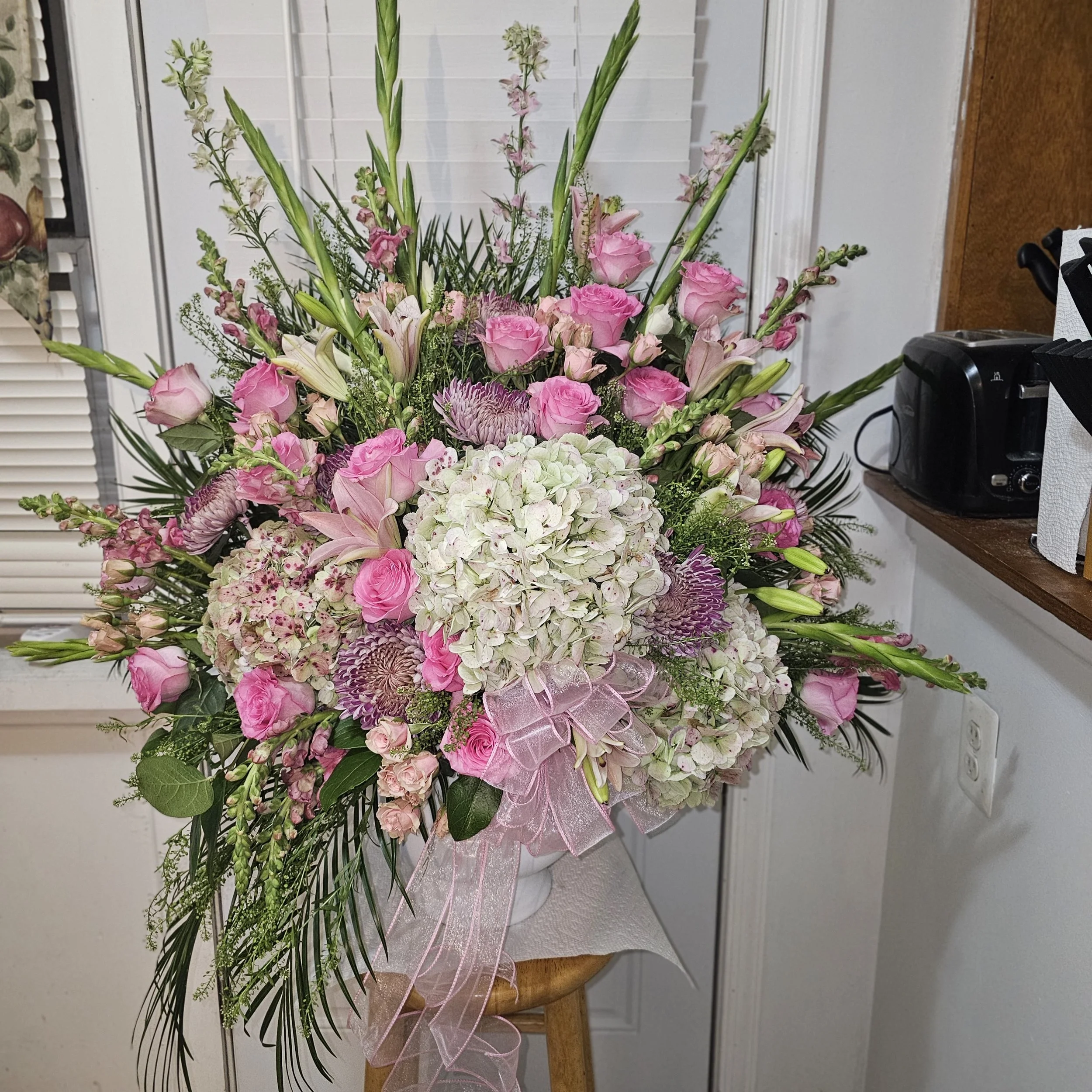 A large floral arrangement featuring pink roses, white hydrangeas, and green foliage, with a pink ribbon bow, set on a wooden table in front of a white wall and window blinds.