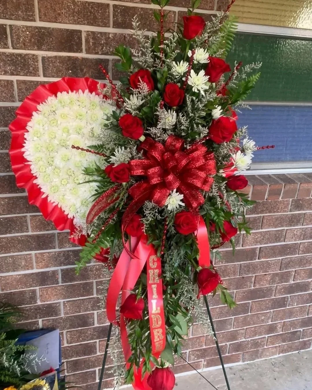Funeral floral arrangement with red roses, white chrysanthemums, greenery, and a large red ribbon with a 'ENDED' message, placed in front of a brick wall.