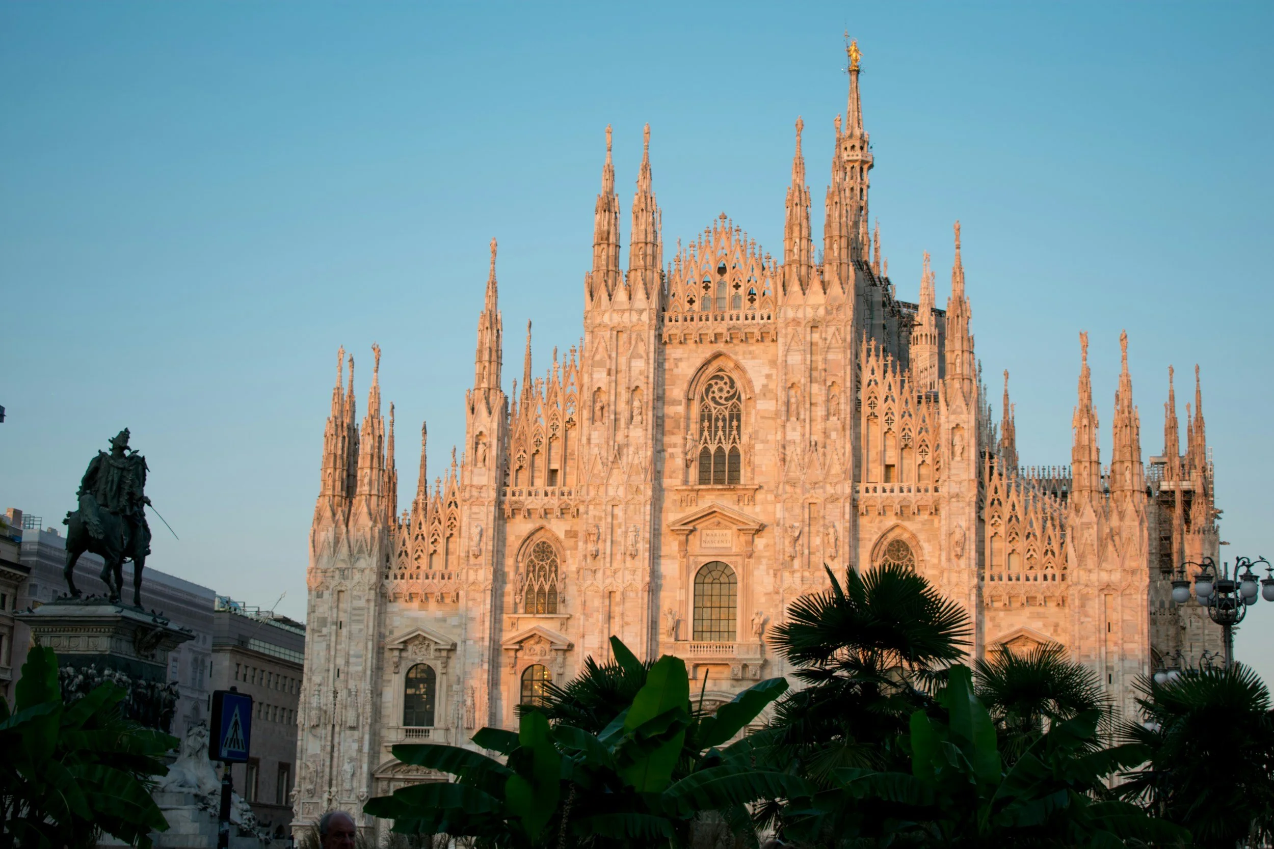 The image shows the Milan Cathedral (Duomo di Milano), a large Gothic cathedral with intricate spires and towers rising into a clear blue sky. In the foreground, there are green plants and a silhouette of a statue of a man on horseback.