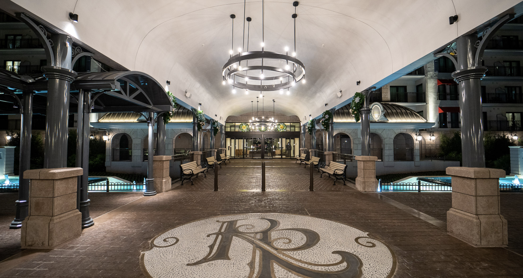 Night view of a covered outdoor walkway in a hotel entrance, decorated with hanging circular light fixtures and a monogrammed design on the floor.