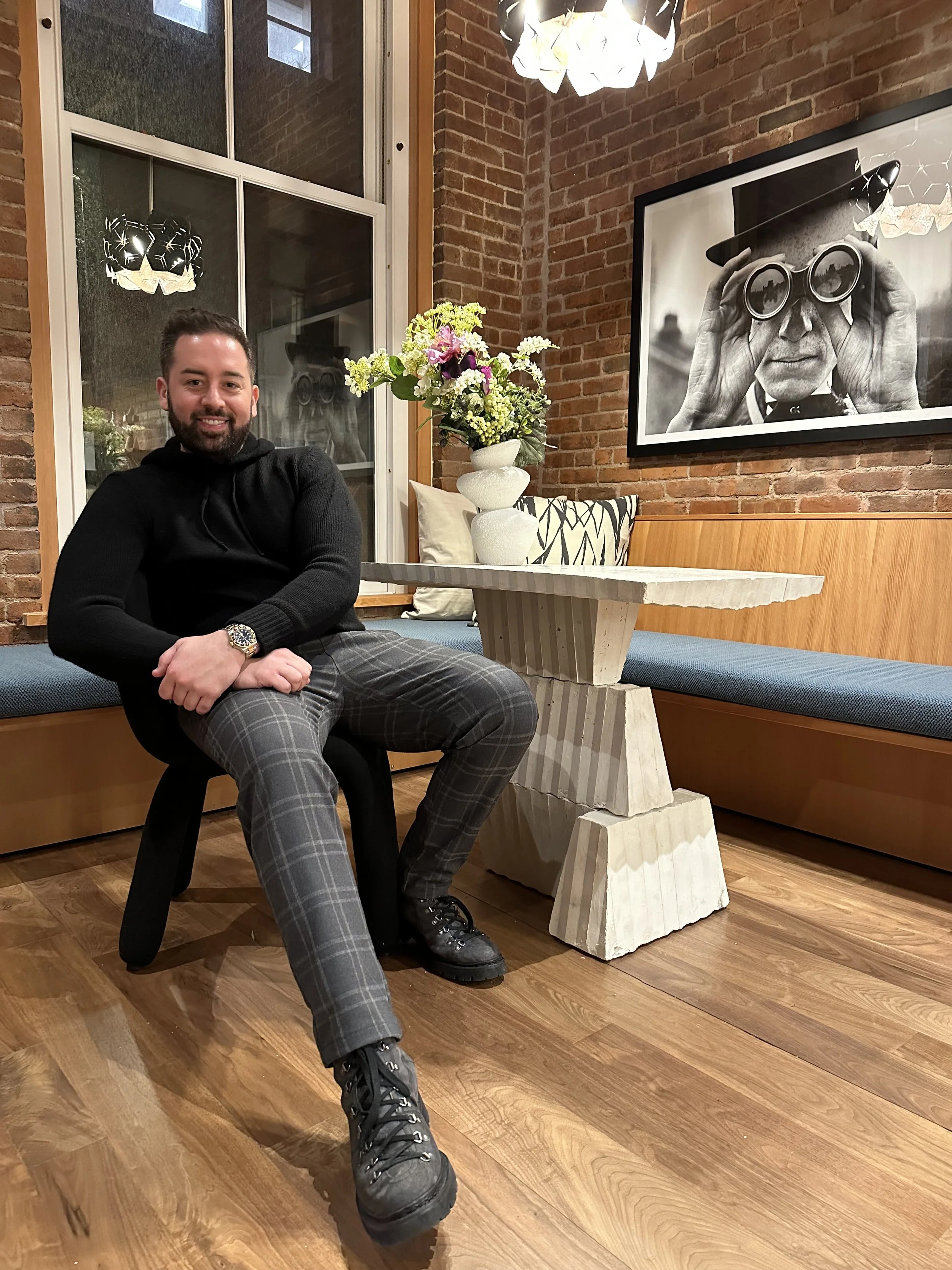 A man sitting on a black chair in a cozy room with a brick wall, large window, and artistic lighting fixture. He is smiling, wearing a black hoodie, plaid pants, and black boots. There is a white stone table with a flower arrangement nearby, and framed black and white photo of a person looking through binoculars hangs on the brick wall.