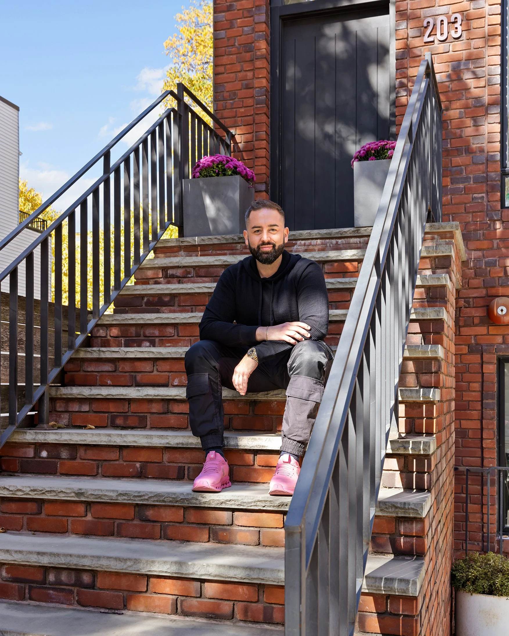 A man with a beard sitting on brick stairs outside a red brick building, wearing a black hoodie, gray and black pants, pink sneakers, with purple flowers in planters beside him.