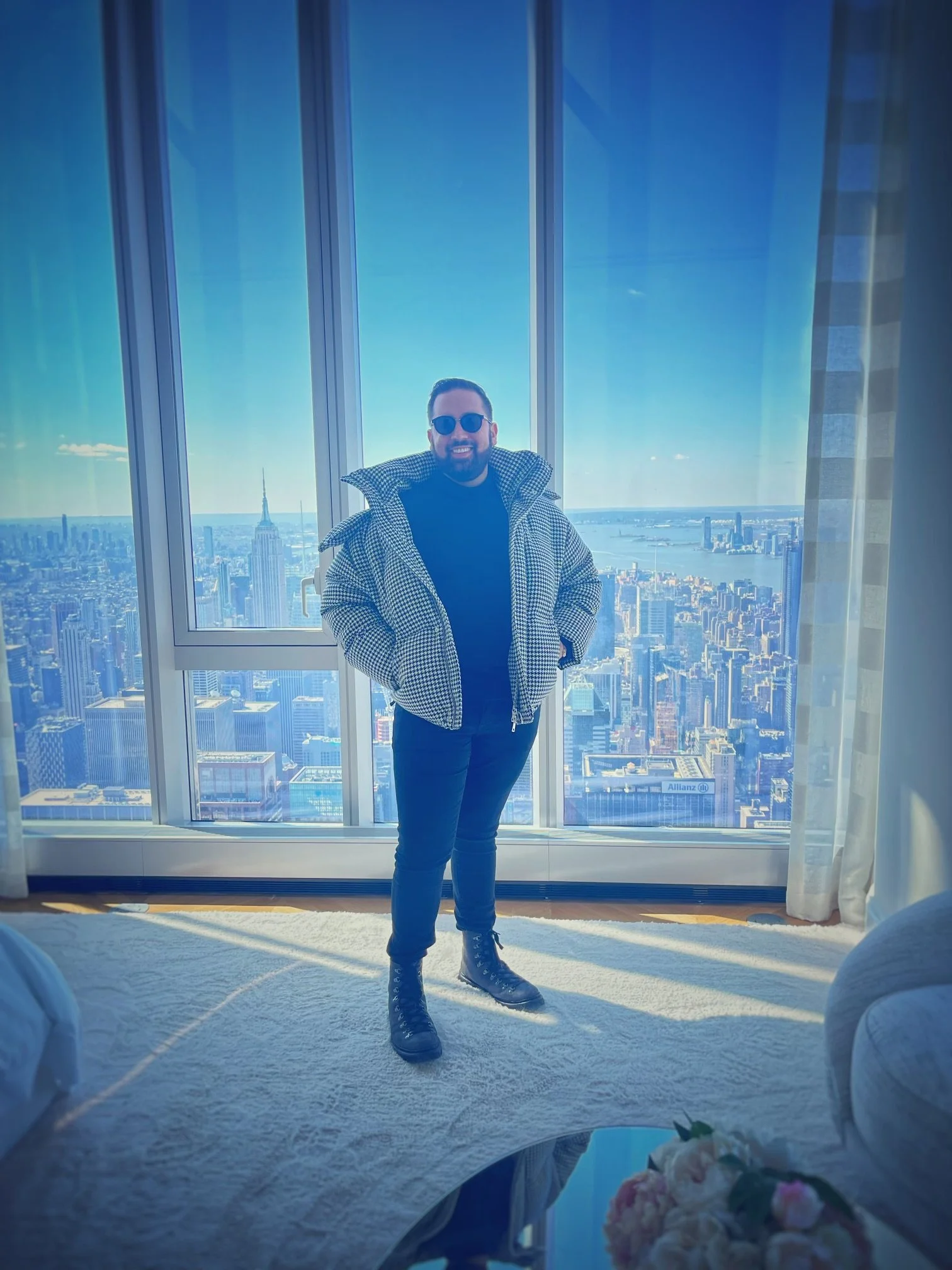 A man standing inside a high-rise apartment with floor-to-ceiling windows showing a city skyline and the Empire State Building, wearing sunglasses, a black shirt, a houndstooth jacket, black pants, and black boots, smiling for the photo.
