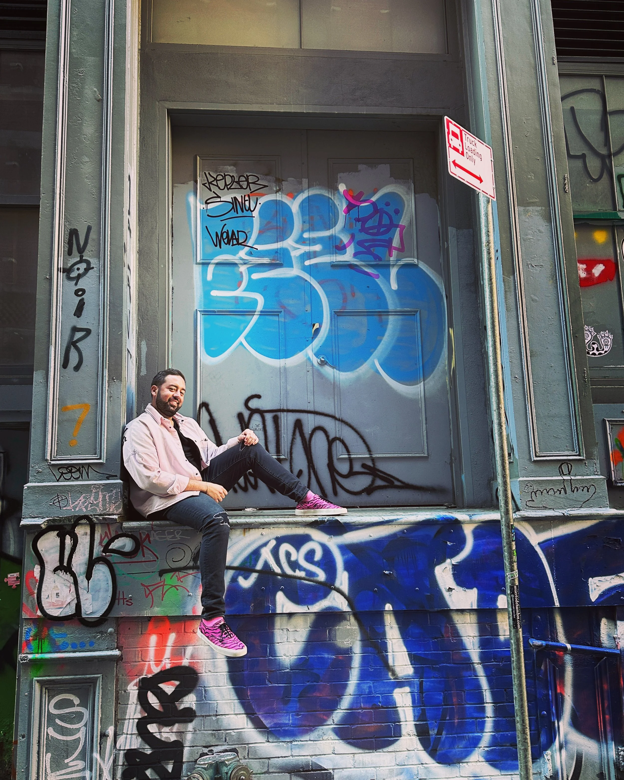 A man with a beard and pink shoes sitting on the ledge of a graffiti-covered building wall, smiling at the camera.