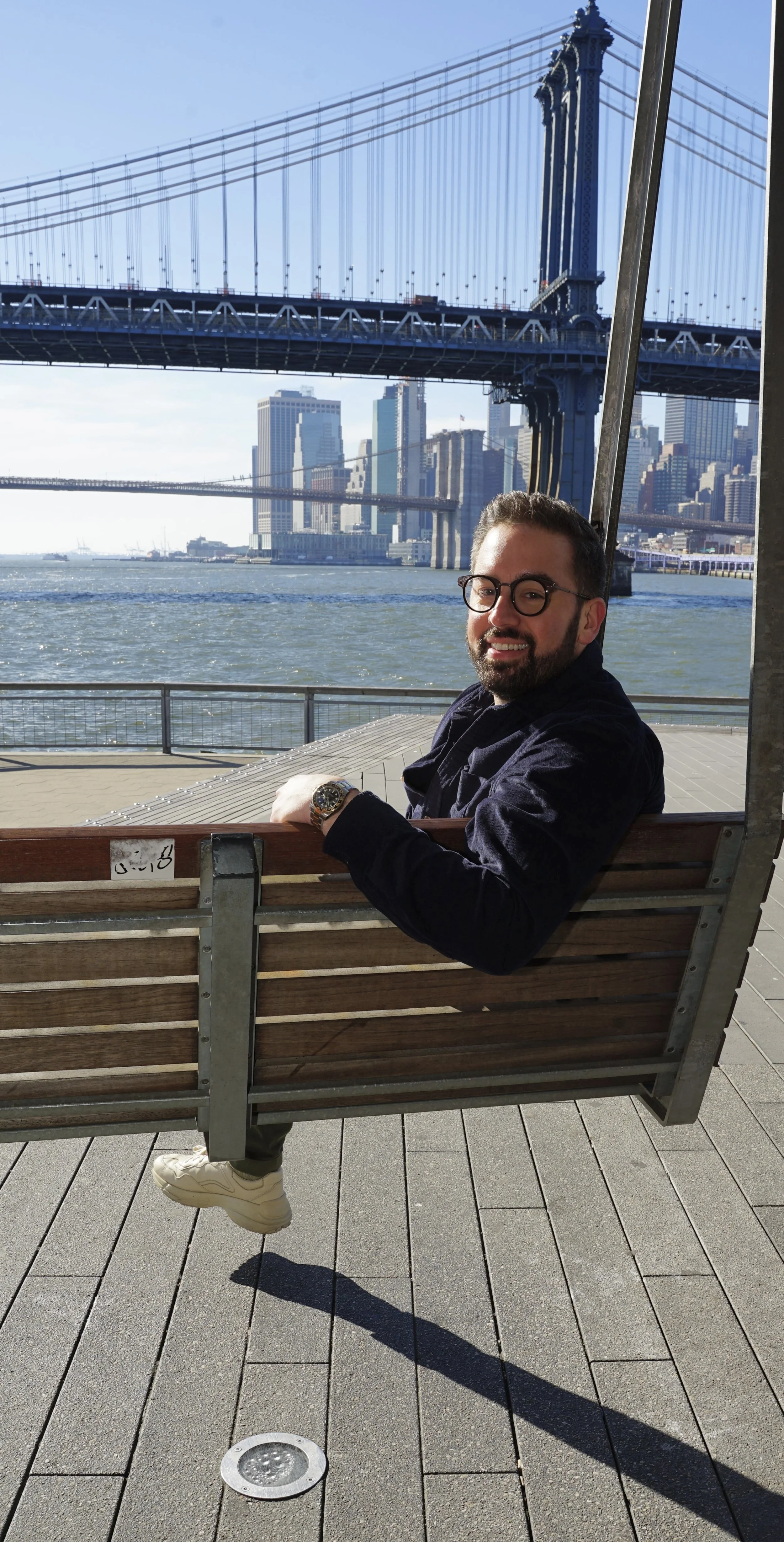 A man sitting on a wooden bench near the waterfront with the Manhattan Bridge and city skyline in the background.