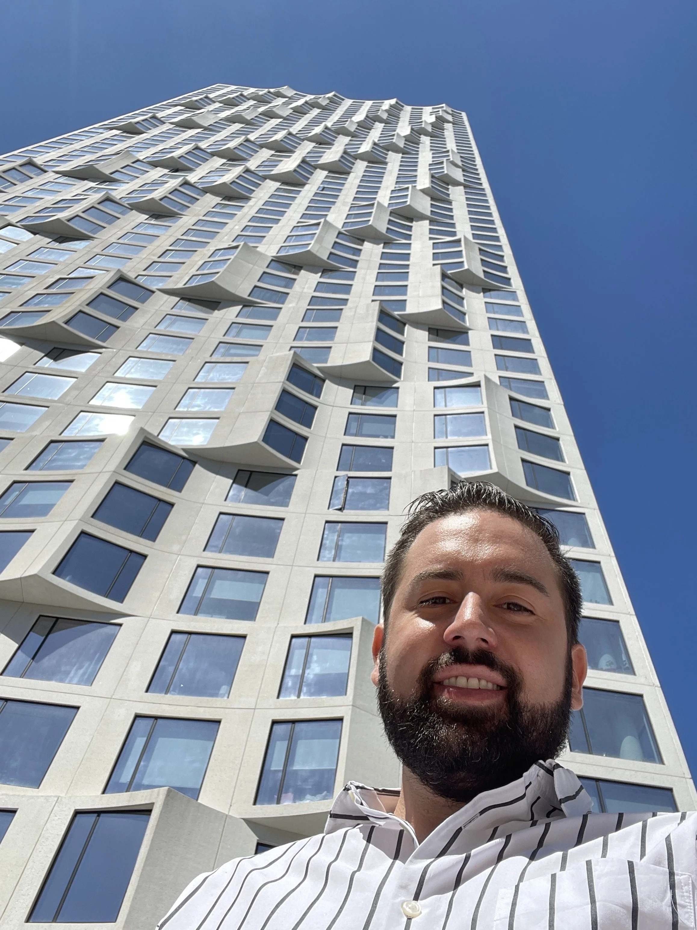 A man with a beard taking a selfie in front of a tall, modern building with unique protruding window structures against a clear blue sky.