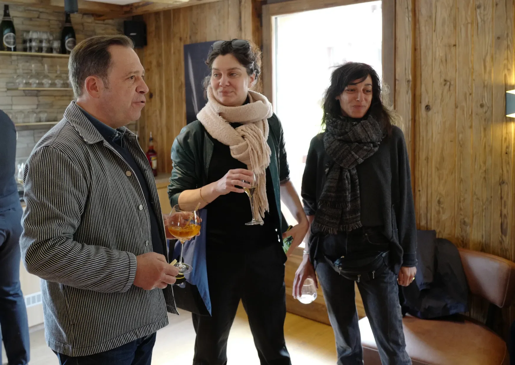 Trois personnes lors d'une réunion ou d'une célébration dans une pièce avec murs en bois, tenant des verres de boissons alcoolisées, deux femmes et un homme, discutant et souriant.