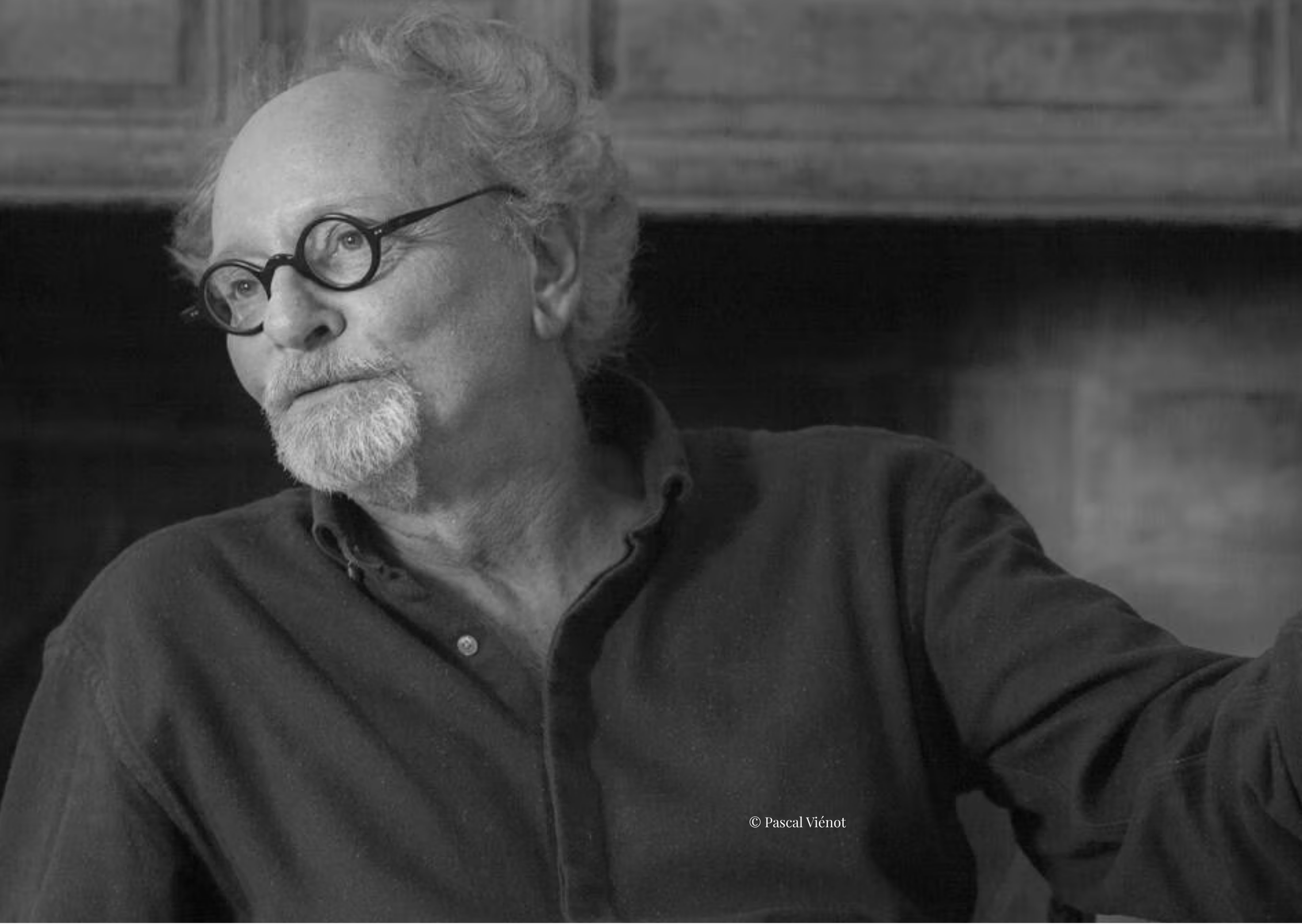 Portrait en noir et blanc d'un homme âgé portant des lunettes rondes, avec une barbe blanche, assis dans un environnement intérieur en bois.