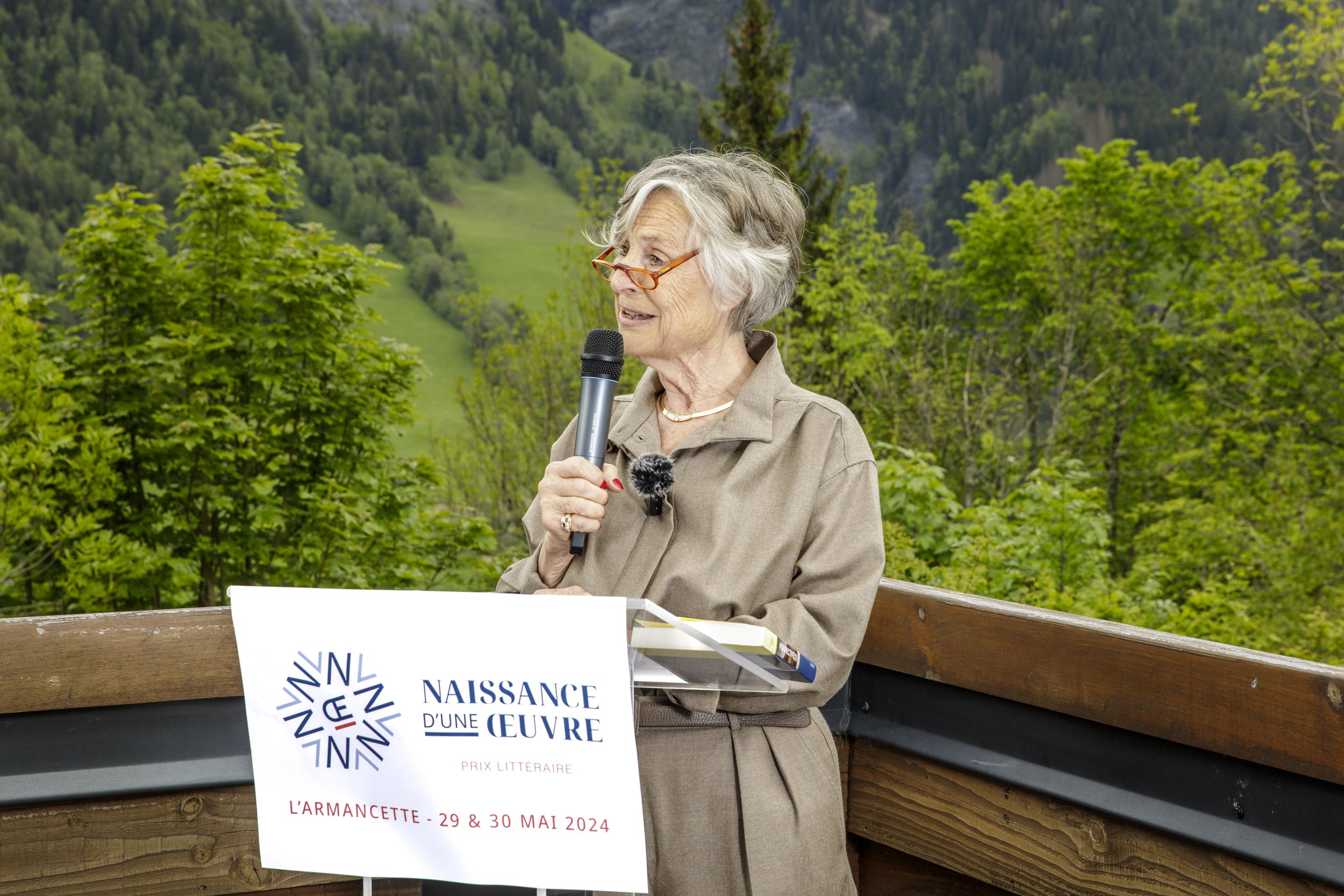 Laurence Viénot tient un micro en parlant devant un panneau indiquant un événement littéraire à L'Armancette, avec un paysage de montagnes et arbres verts en arrière-plan.