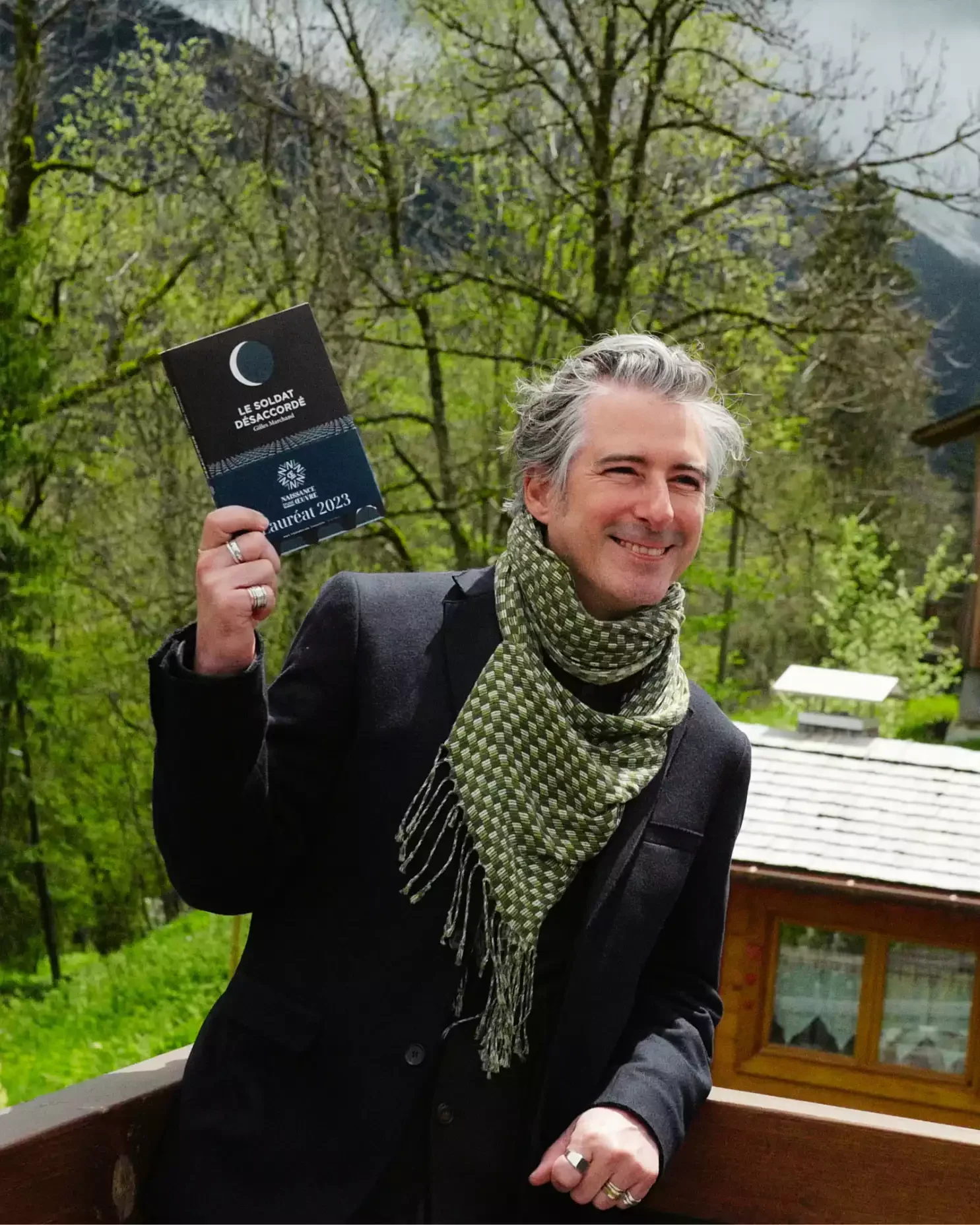 Un homme souriant tient un livre à la main, il porte un costume noir et une écharpe vert foncé à motifs enroulée autour du cou, dans un environnement extérieur verdoyant avec des arbres et une petite construction en bois en arrière-plan.