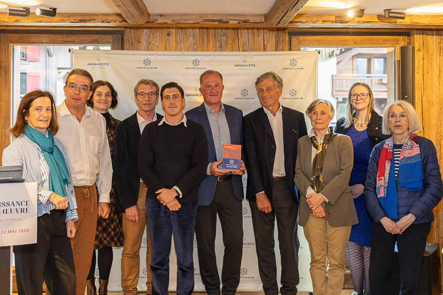 Groupe de personnes posant pour une photo lors d'un événement littéraire. L'arrière-plan affiche un panneau avec le texte "Naissance de l'Œuvre" et la date 22 mai 2025.