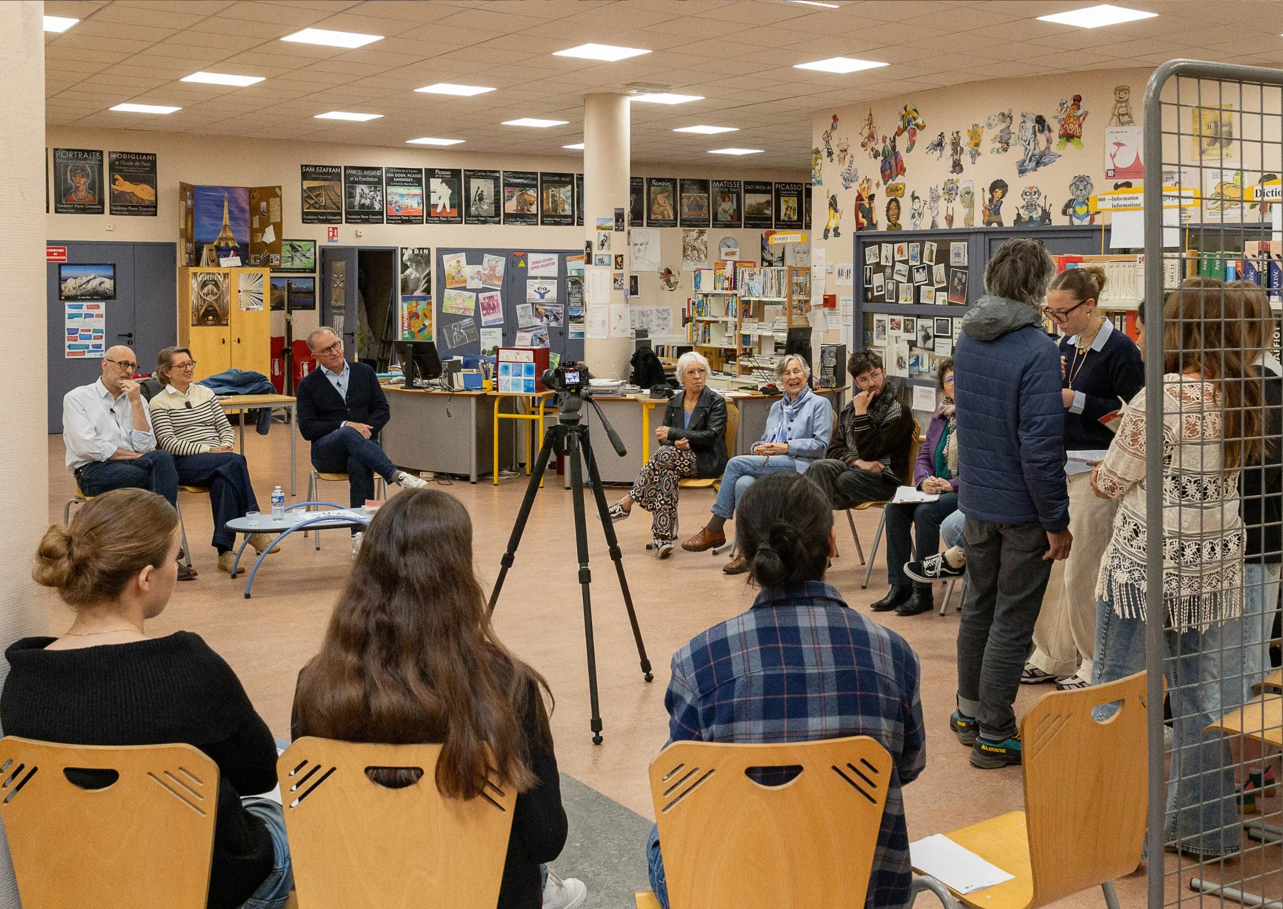 Groupe de personnes assises et discutant dans une bibliothèque ou un centre communautaire, avec deux femmes debout en train de parler. Mur remplis d'affiches, livres et œuvres d'art décorent la pièce.