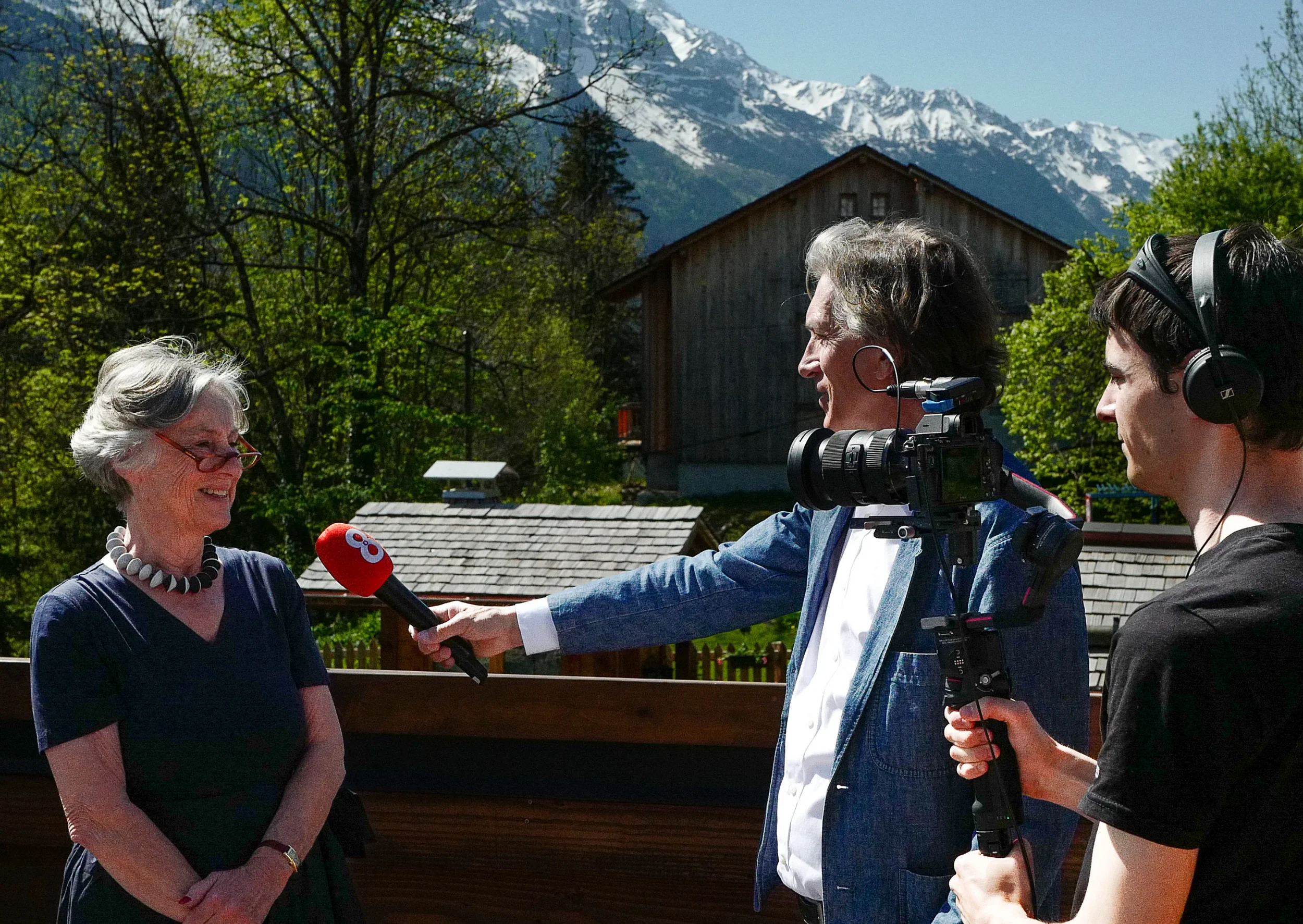 Une interview est en cours avec une femme âgée souriante, interviewée par un journaliste avec une caméra et une perche microphone rouge, au fond de montagnes enneigées et d'arbres verts.