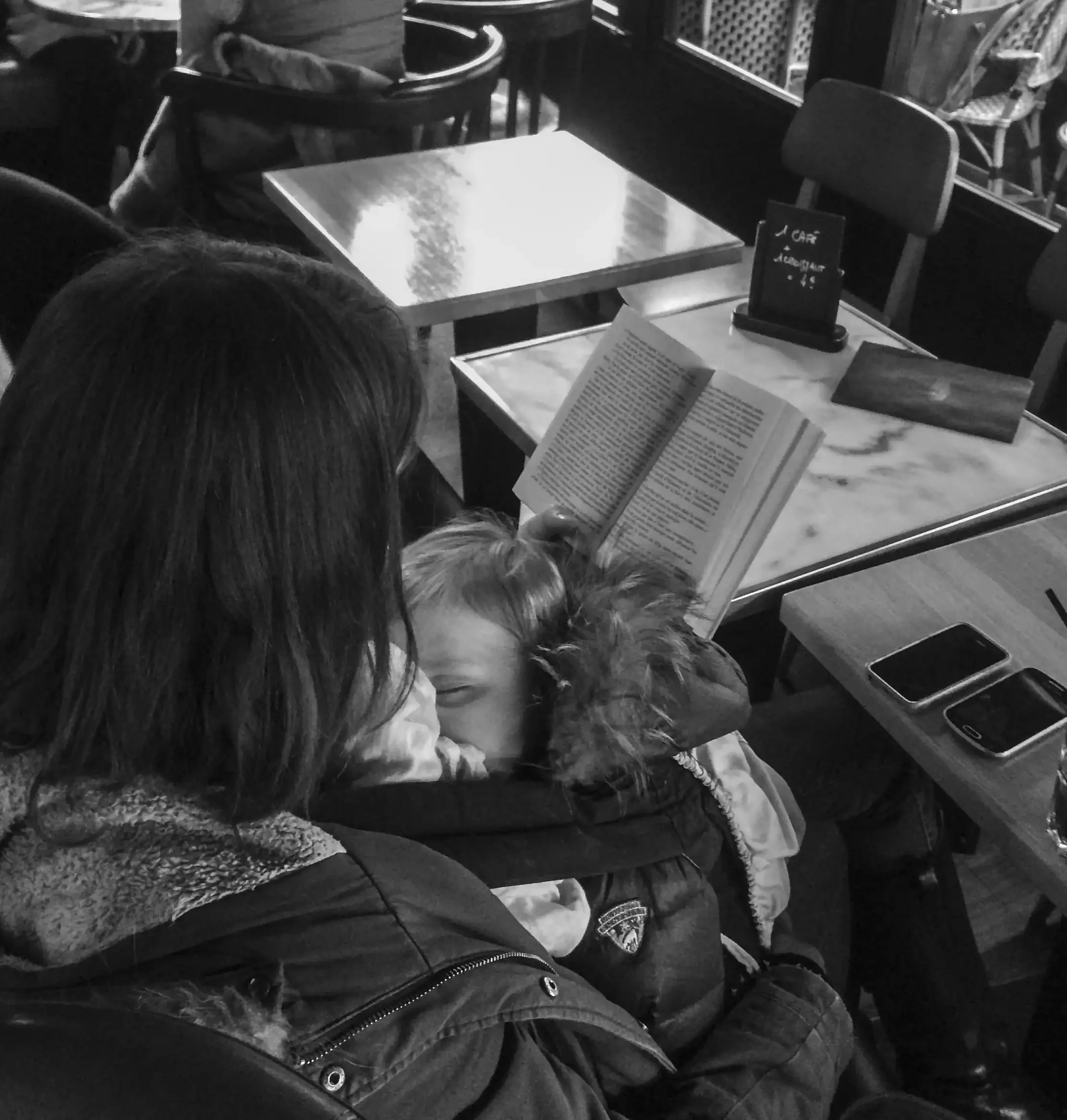 Deux personnes assises à une table dans un café, lisant un menu ou un livre. L'ambiance est décontractée. La photo est en noir et blanc.