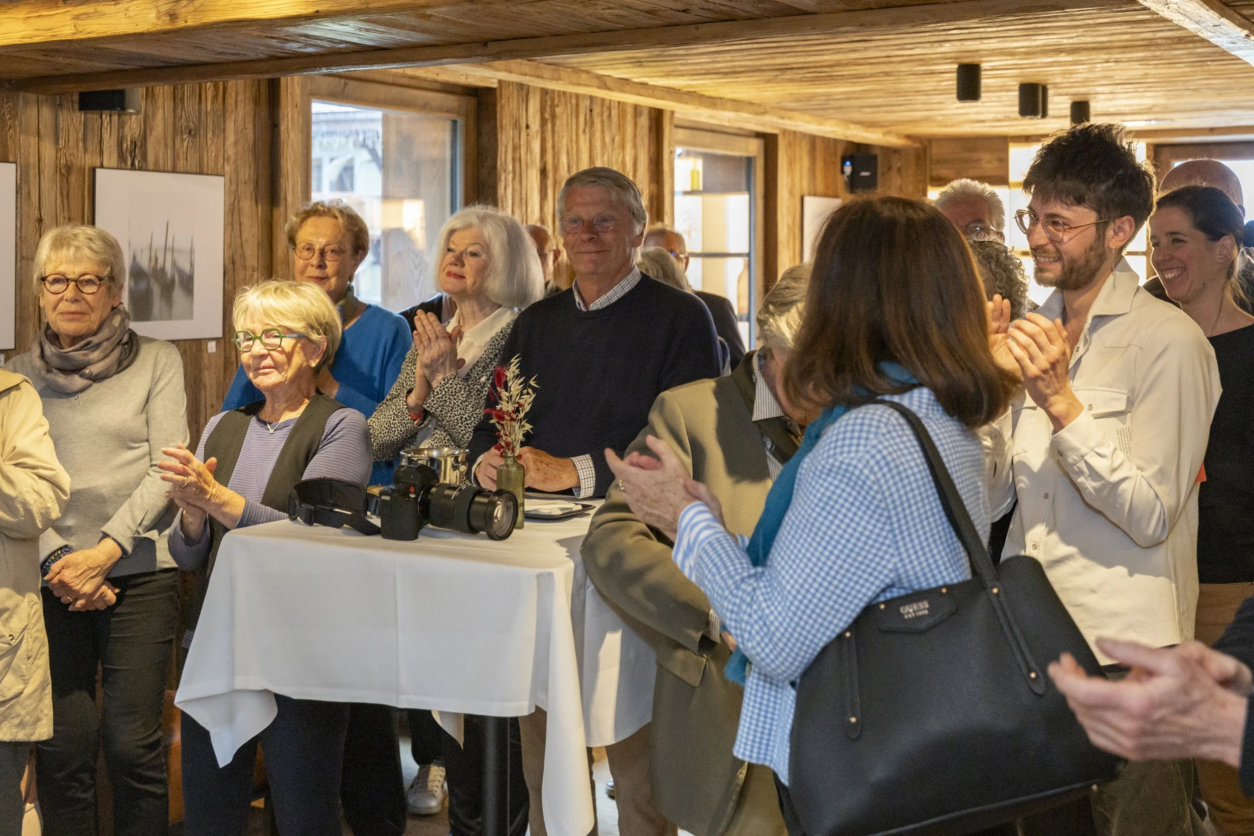 Groupe de personnes souriantes et applaudissant lors d'un événement à l'intérieur, avec des murs en bois et des fenêtres derrière eux.