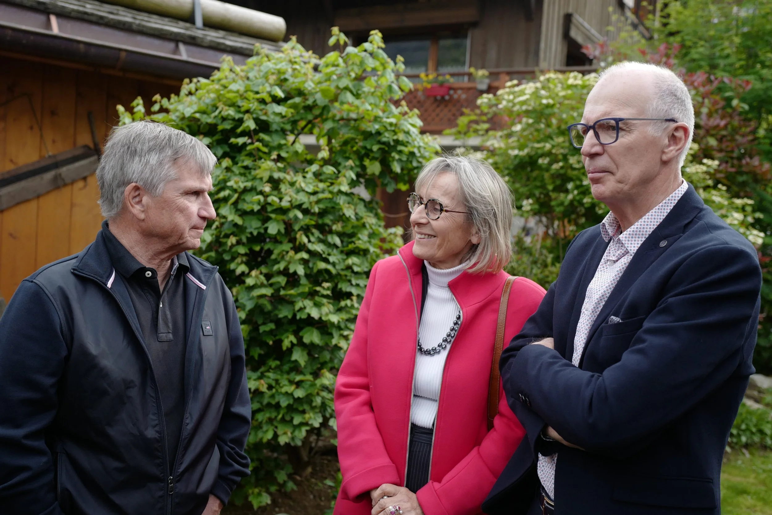 Trois personnes discutent dans un jardin. Deux hommes et une femme, tous portant des vêtements formels. Les hommes portent des lunettes. La femme porte un blazer rose.