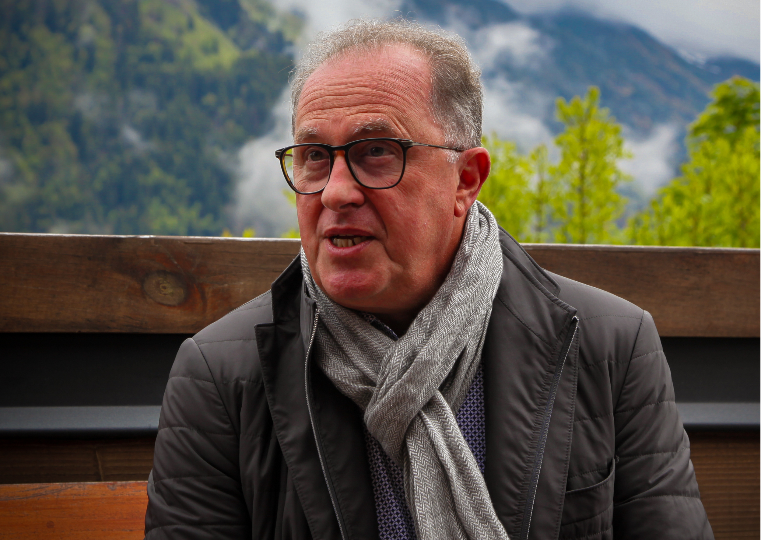 Laurent Seyer, en veste, assis sur un banc en bois, avec un paysage de montagnes et de forêts en arrière-plan.